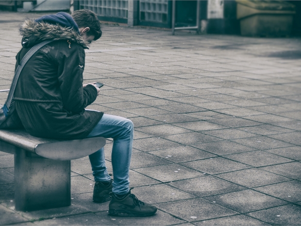 image of a man sitting ona  bench in a coat faced away from the camera while looking down on his phone